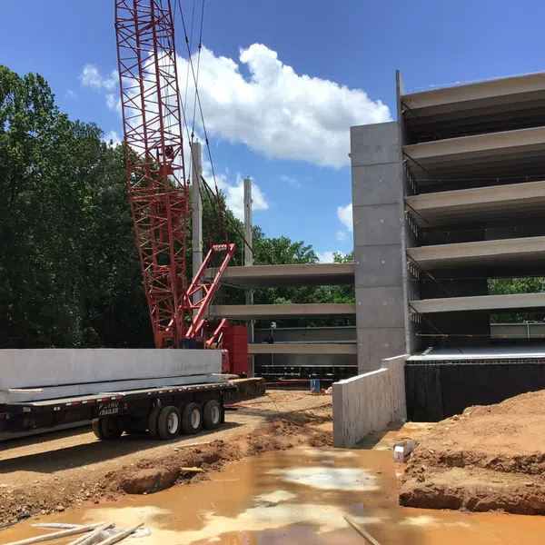 Construction site with crane, multi-story concrete parking structure, red dirt, and workers installing precast concrete beams
