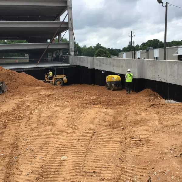 Construction site with workers operating machinery and compacted dirt near a multi-level concrete structure.