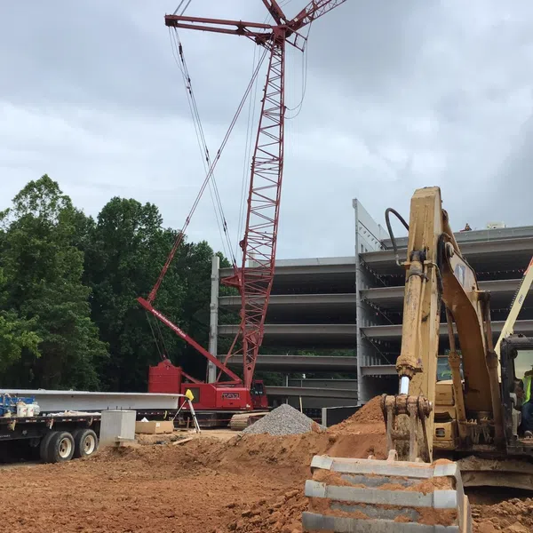 Construction site with excavator, crane, and a multi-story concrete building under construction surrounded by trees.