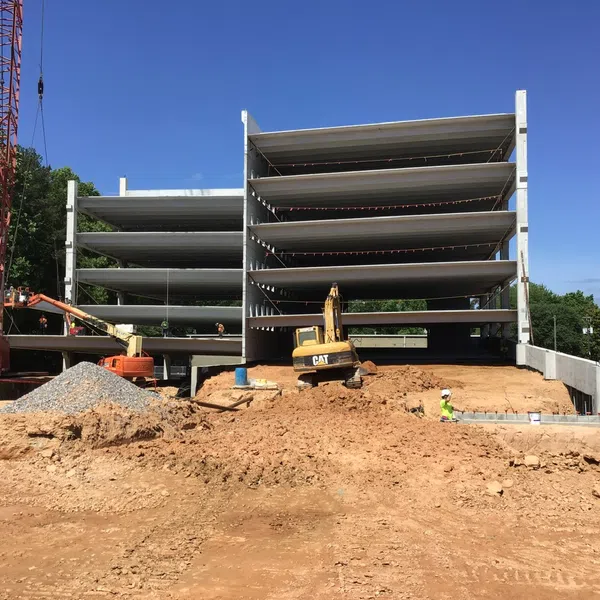 Multi-level concrete parking garage under construction with cranes, excavator, and workers on dirt site under clear sky