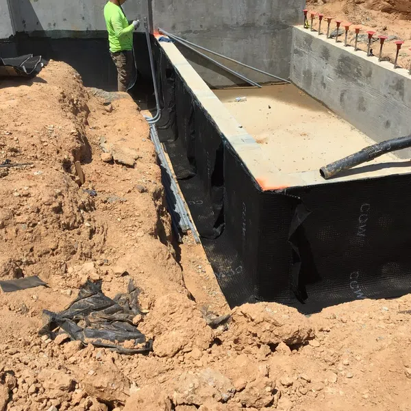 Construction site with foundation waterproofing being applied and workers installing drainage pipes around a building base.