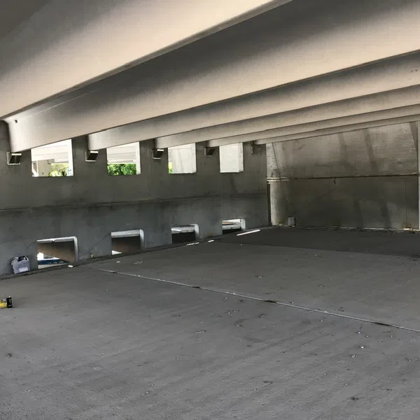 Empty concrete parking garage level with beams, open side windows, and scattered debris on floor under daylight.