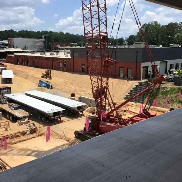 Construction site with crane unloading concrete slabs, trailers, and industrial buildings under clear blue sky.