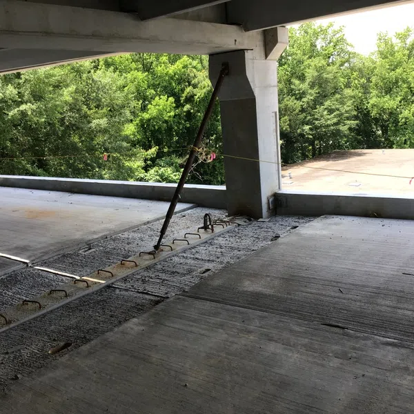 Under construction concrete parking garage floor with exposed rebar and support beams, surrounded by green trees.