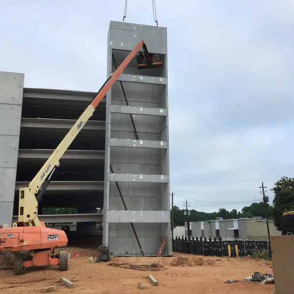 Construction site with a boom lift working on a tall concrete structure beside a multi-level parking garage under cloudy sky.