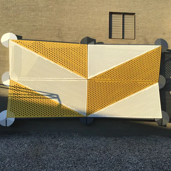 Geometric yellow and white perforated metal panels mounted on beige brick wall casting shadows.