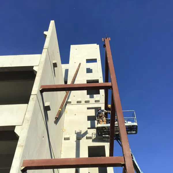 Construction site with workers on a lift installing steel beams against a concrete building under clear blue sky