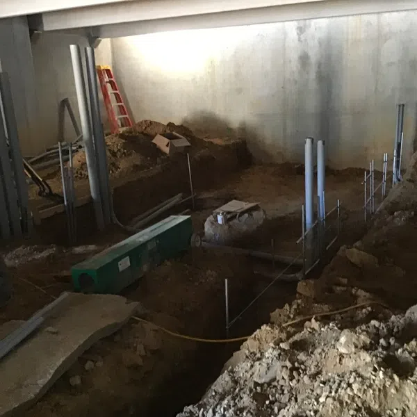 Construction site showing foundation work with exposed dirt, pipes, and a worker in a green shirt inside a building.