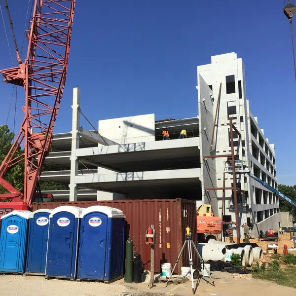 Construction site with concrete building structure, red crane, blue portable toilets, and workers under clear sky.