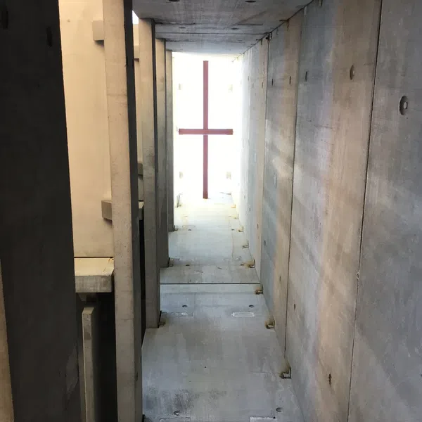 Tall concrete corridor leading to a glowing cross-shaped window at the far end with light streaming through.