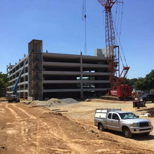 Construction site with a multi-level concrete parking structure, crane, pickup trucks, and dirt ground under clear blue sky