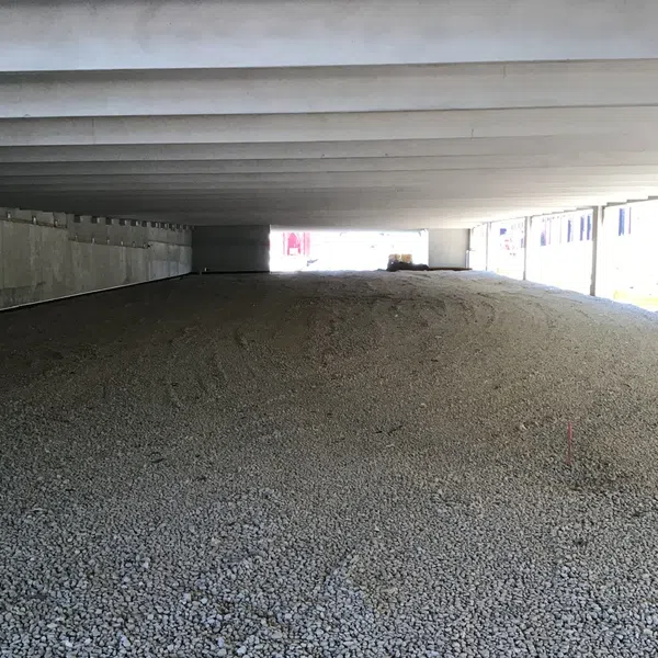 Underneath a concrete bridge with beams, a large pile of gravel covers the ground, illuminated by natural light.