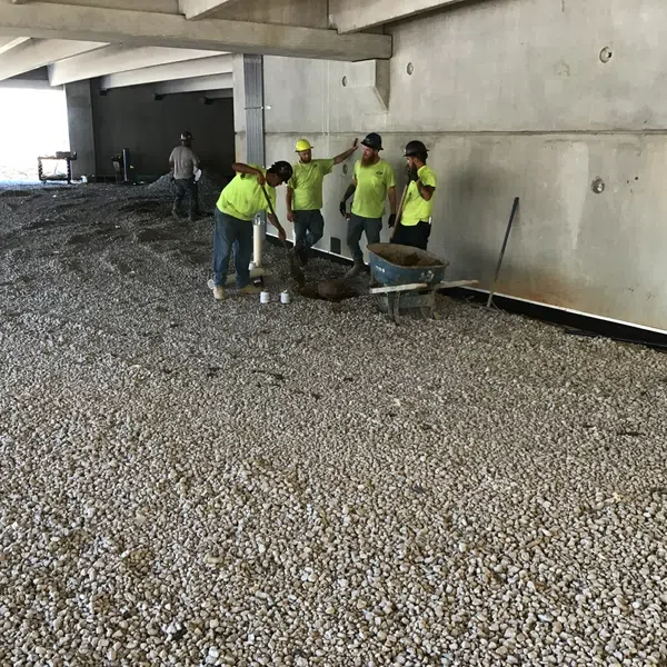 Construction workers in yellow shirts spreading gravel under a concrete structure at a building site.