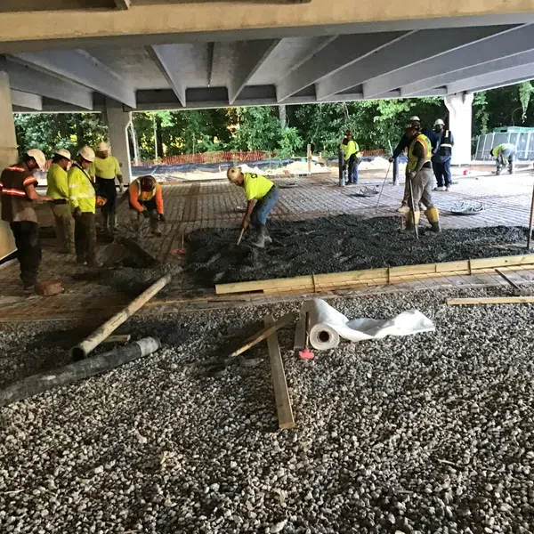 Construction workers pouring and leveling concrete under a highway overpass during daytime.