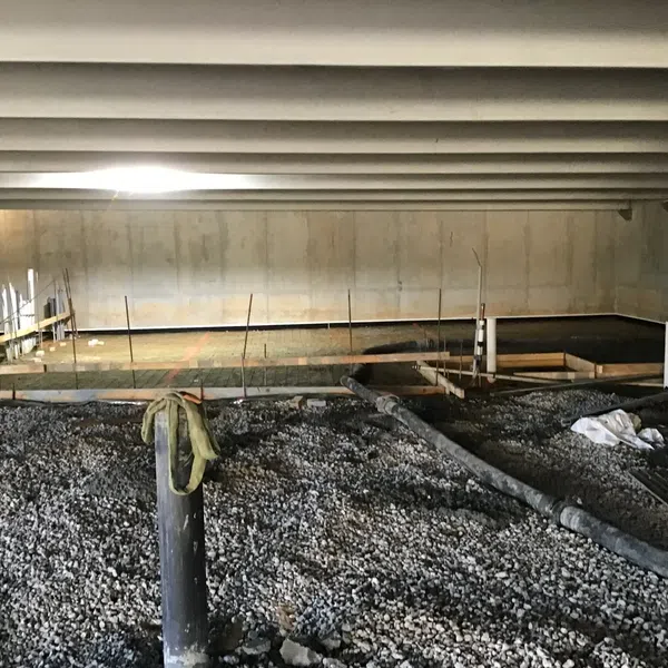 Construction worker overseeing gravel and concrete foundation work in an indoor building site with exposed ceiling beams.