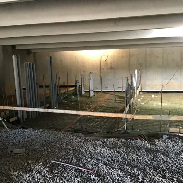 Indoor construction site with gravel floor, plumbing pipes, and concrete slab preparation under beams.