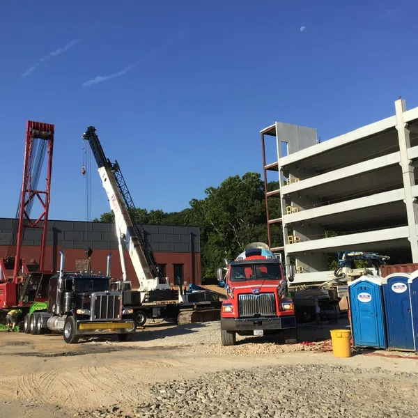 Construction site with cranes, trucks, portable toilets, and a partially built multi-story parking garage under clear blue sky.