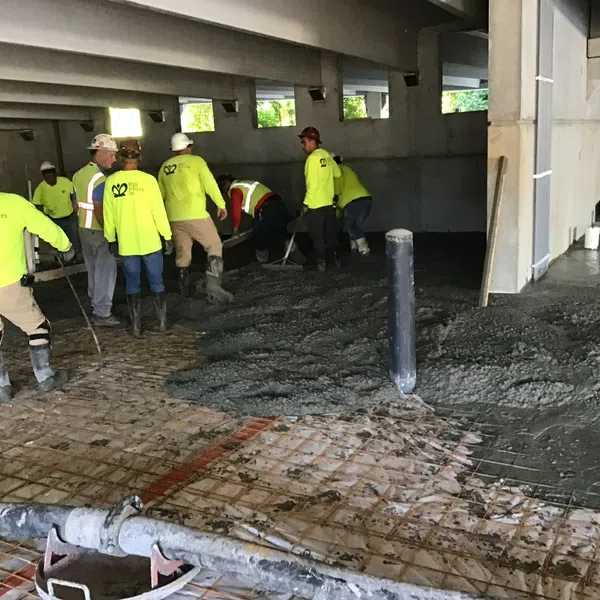 Construction workers in safety gear pour and spread concrete inside a building under construction with exposed beams.