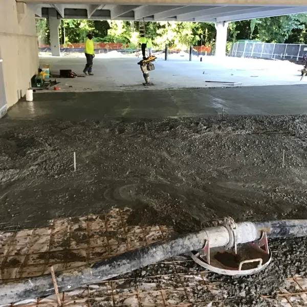 Construction workers smoothing freshly poured concrete on a large outdoor slab under a shaded structure.