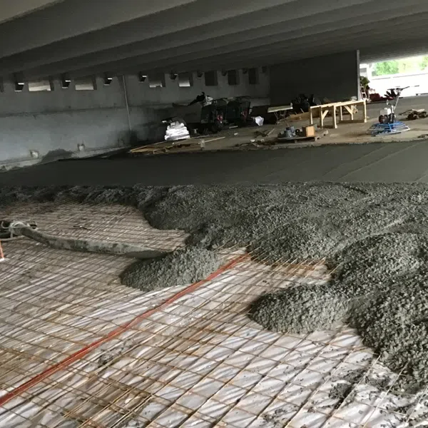 Concrete being poured and leveled over steel rebar mesh reinforcement at a construction site under a bridge.