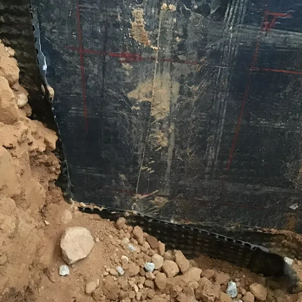 Close-up of basement foundation wall with black waterproofing membrane and loose soil with small rocks at base.