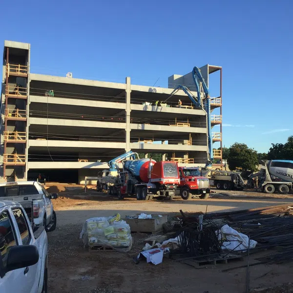 Concrete parking garage under construction with cement trucks, construction vehicles, and workers at site under clear sky.