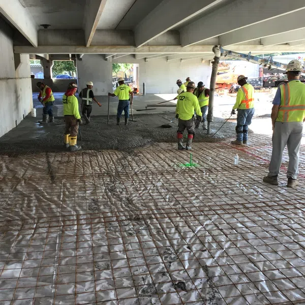 Construction workers pouring and leveling concrete under a bridge on a reinforced wire mesh base.