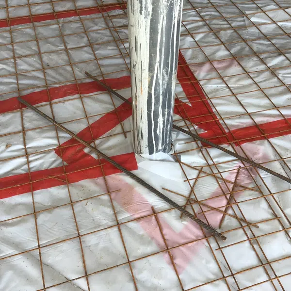 Concrete reinforcement grid with rusted rebar and protective metal column in construction site covered in plastic sheeting.