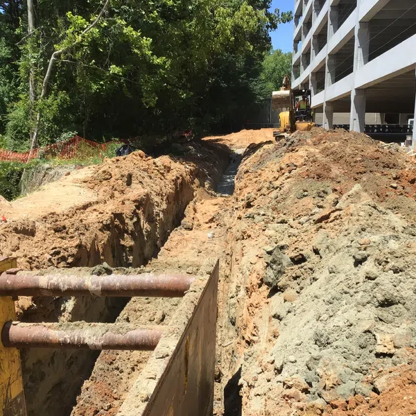 Deep trench excavation with metal supports near a large building under construction surrounded by trees and soil piles.
