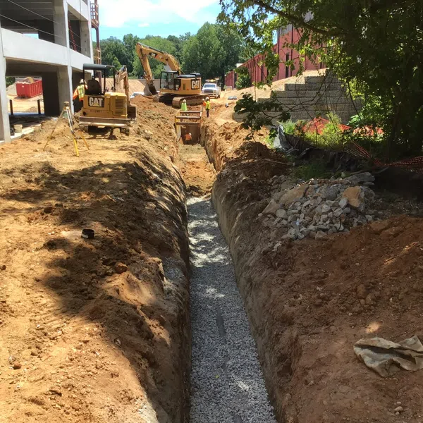 Deep trench filled with gravel at a construction site with excavators and building structures nearby under blue sky.