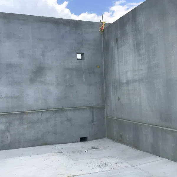 Empty gray concrete room with high walls under blue sky with white clouds, featuring small wall openings and construction markings.
