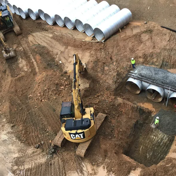 Excavators working on large trench with stacked metal pipes and workers installing drainage system