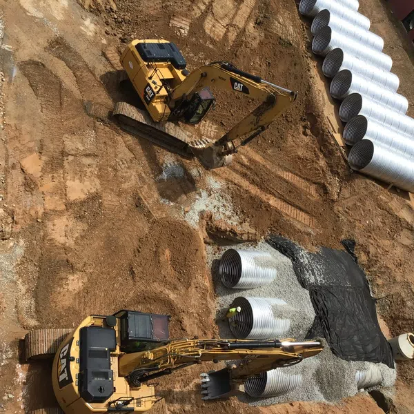 Aerial view of construction site with two yellow excavators and large concrete drainage pipes on dirt ground.