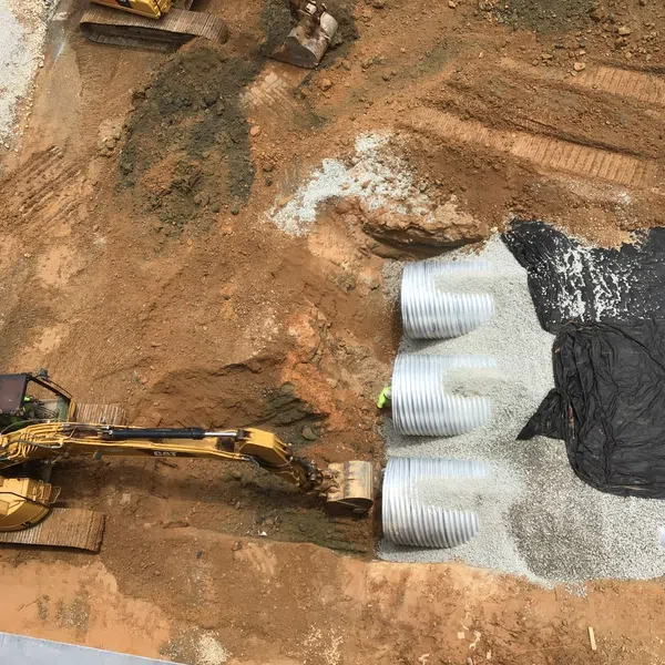 Construction site with excavator digging soil near large corrugated pipes and gravel bedding.