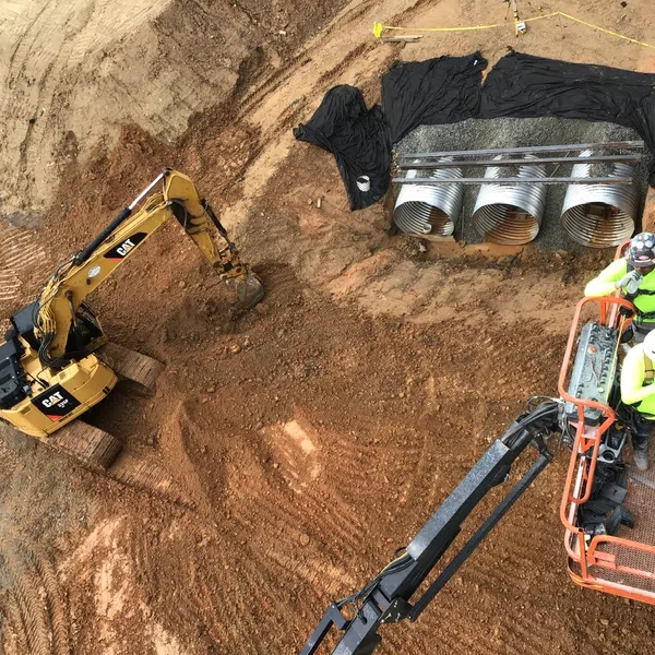 Aerial view of construction site with excavator, workers in lift, and large drainage pipes on dirt ground.