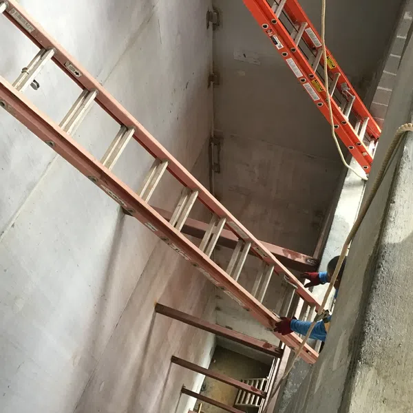 Worker setting up long ladders inside a deep concrete construction shaft with safety gear.