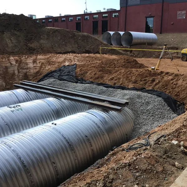 Construction site with large metal drainage pipes being installed in an excavated trench beside a building.