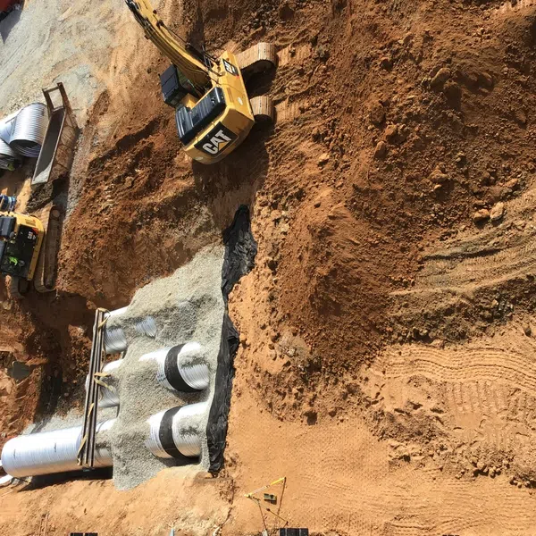 Construction site with excavators digging and large metal pipes being installed underground in dirt and gravel.
