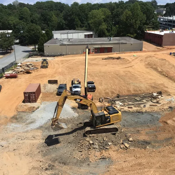 Construction site with excavator moving earth, surrounded by equipment, vehicles, and buildings in a commercial area.