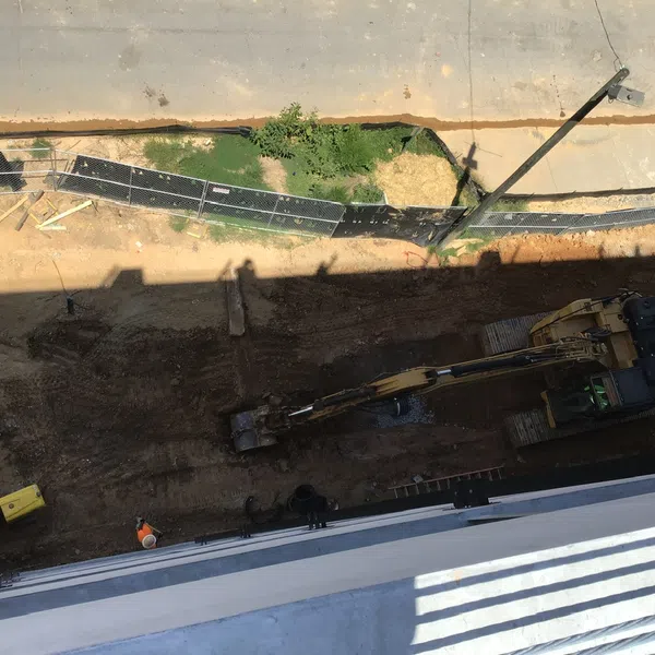Aerial view of construction site with excavator, dirt ground, fencing, and a parked white vehicle on paved road.