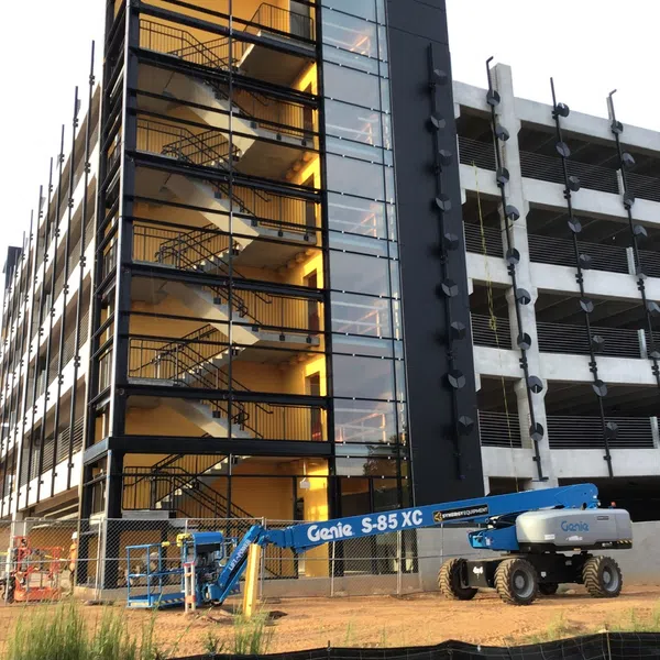 Construction site with modern parking garage featuring glass stairwell and blue Genie S-85 XC lift machine.