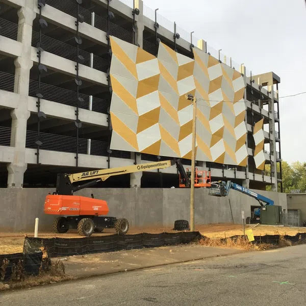 Construction site with lifts installing large yellow and white geometric panels on a multi-story parking structure.