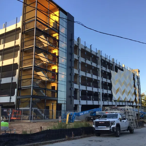 Under-construction multi-level parking garage with yellow stairwell lighting and construction vehicles outside at dusk