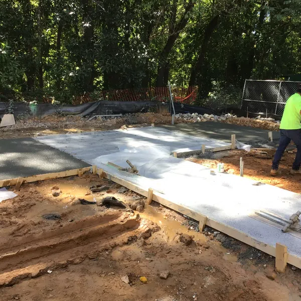 Construction worker smoothing freshly poured concrete slabs on a cleared dirt site surrounded by trees.