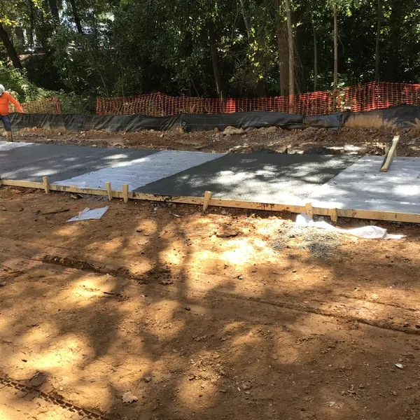 Worker smoothing freshly poured concrete slab at outdoor construction site surrounded by trees and orange safety fencing.