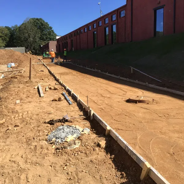 Construction site with workers preparing a long concrete foundation next to a red brick building under clear blue sky.