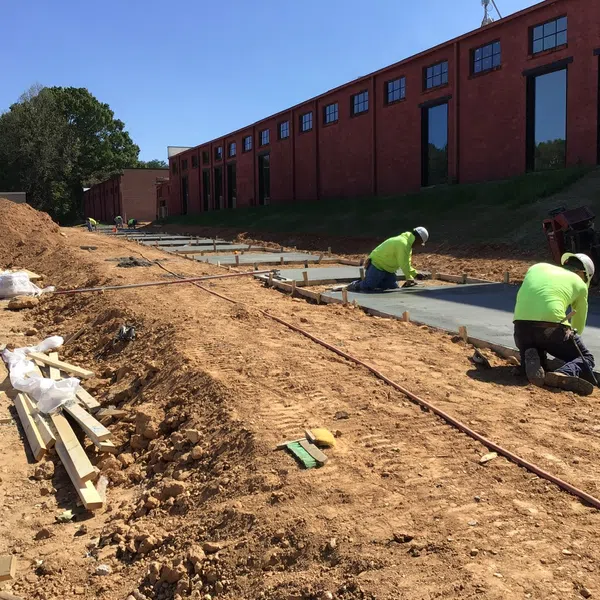 Two construction workers in neon shirts smoothing newly poured concrete sidewalk next to a large red brick building on a sunny day.