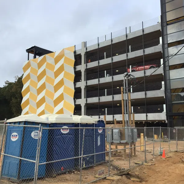 Multi-level parking garage under construction with geometric yellow mural and temporary portable toilets fenced off