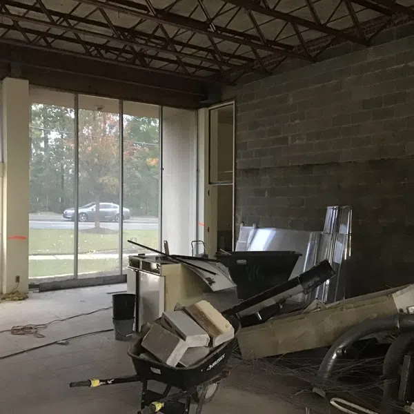 Construction site interior with exposed ceiling, wheelbarrow filled with debris, and large windows showing outside.