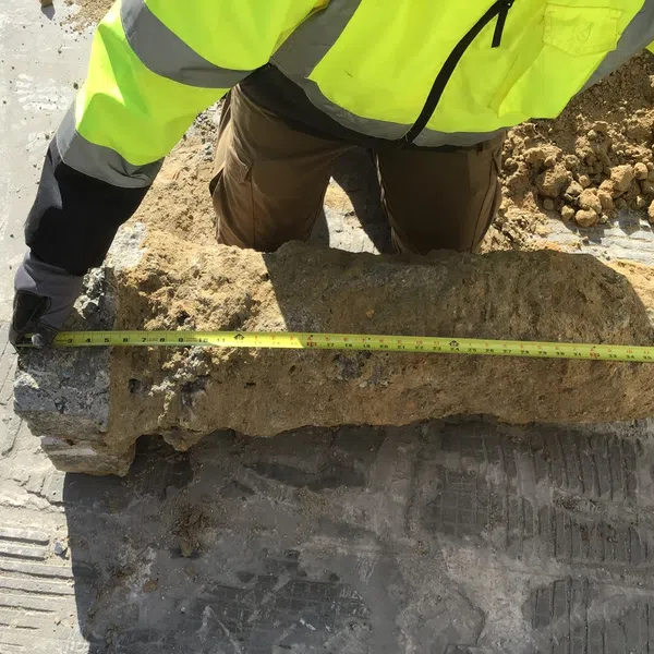 Worker in high-visibility jacket measuring an ancient stone artifact partially covered in dirt on a construction site.